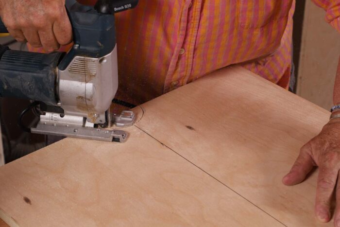 Beth uses a jigsaw to cut out a half circle on a piece of plywood. 